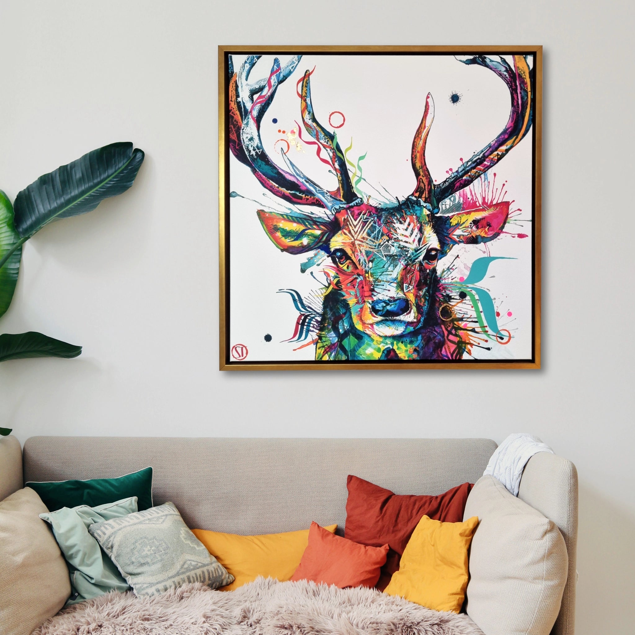 Artist embellished colourful deer artwork with gold frame on a wall above a sofa with colourful cushions.